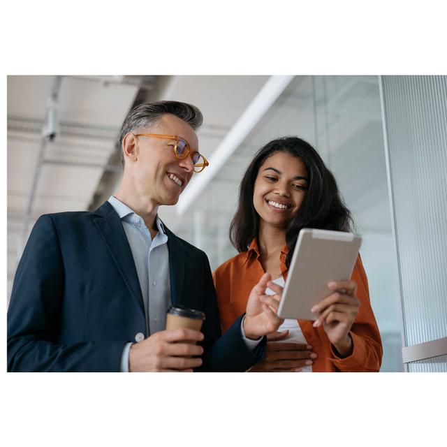 Two fashionable colleagues, a man and a woman, smile as they consult something displayed on a tablet that is held by the woman in an office setting. The man holds a coffee cup.