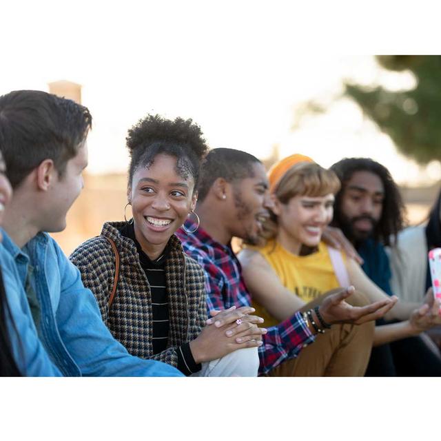 A diverse group of men and women sit together outside, laughing smiling and talking.