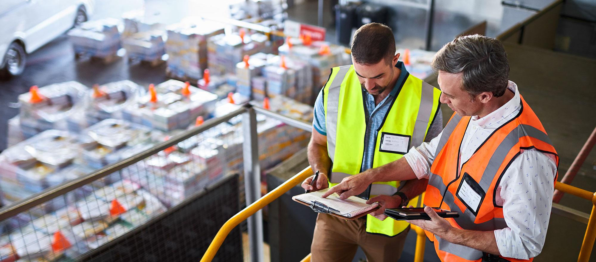 Two men consult a clipboard in a warehouse setting. They are wearing safety vests.