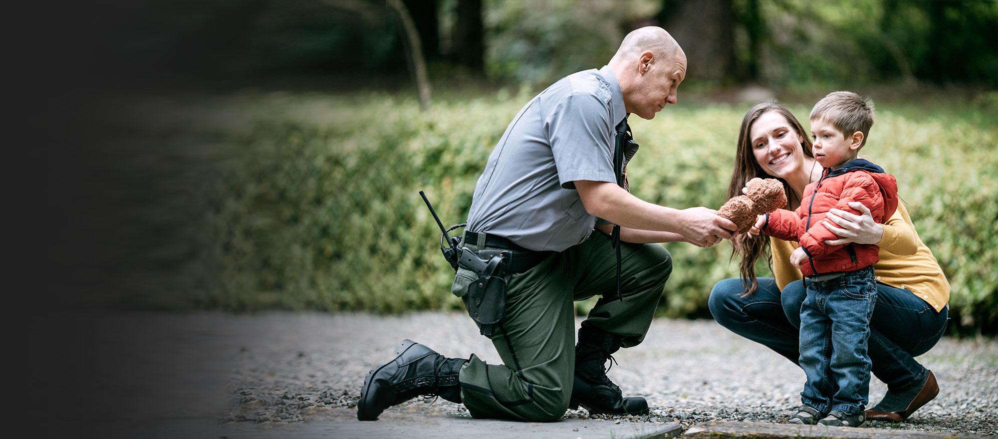 A male public safety law enforcement officer kneels down to give a toy to a child, who is being hugged by his smiling mother.