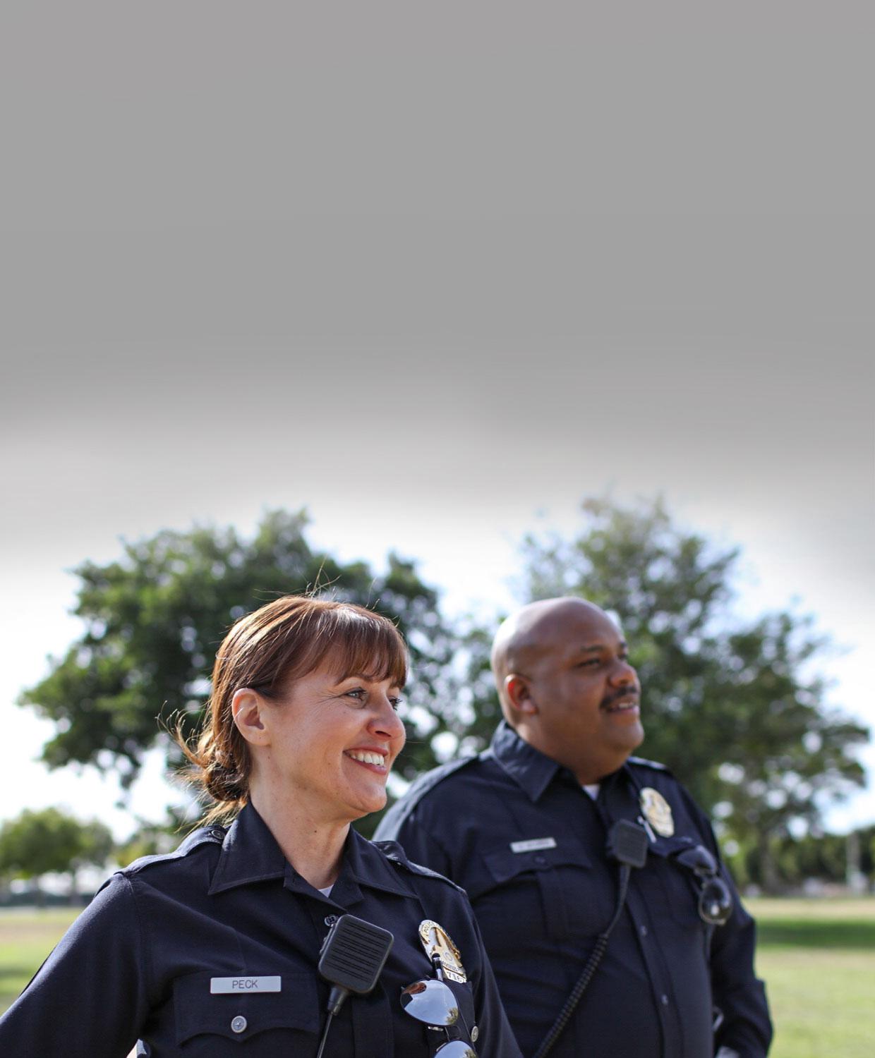 Two public safety law enforcement officers stand in an outdoor setting, smiling off screen.