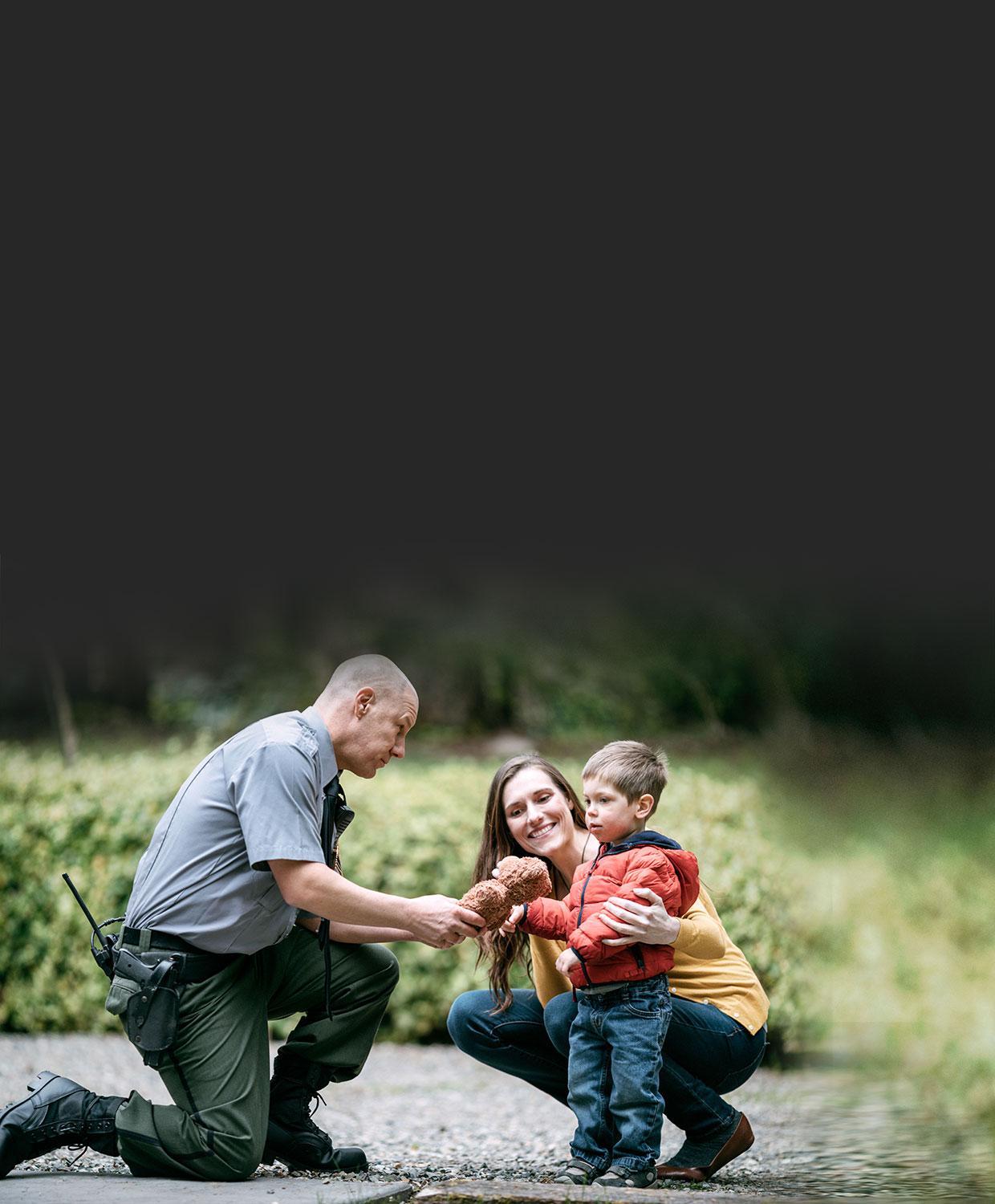 A male public safety law enforcement officer kneels down to give a toy to a child, who is being hugged by his smiling mother.