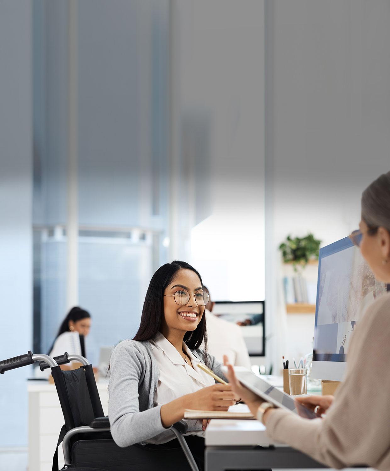 A woman in a wheelchair sits at a desk and smiles as she talks to another woman who holds a tablet in an office setting.