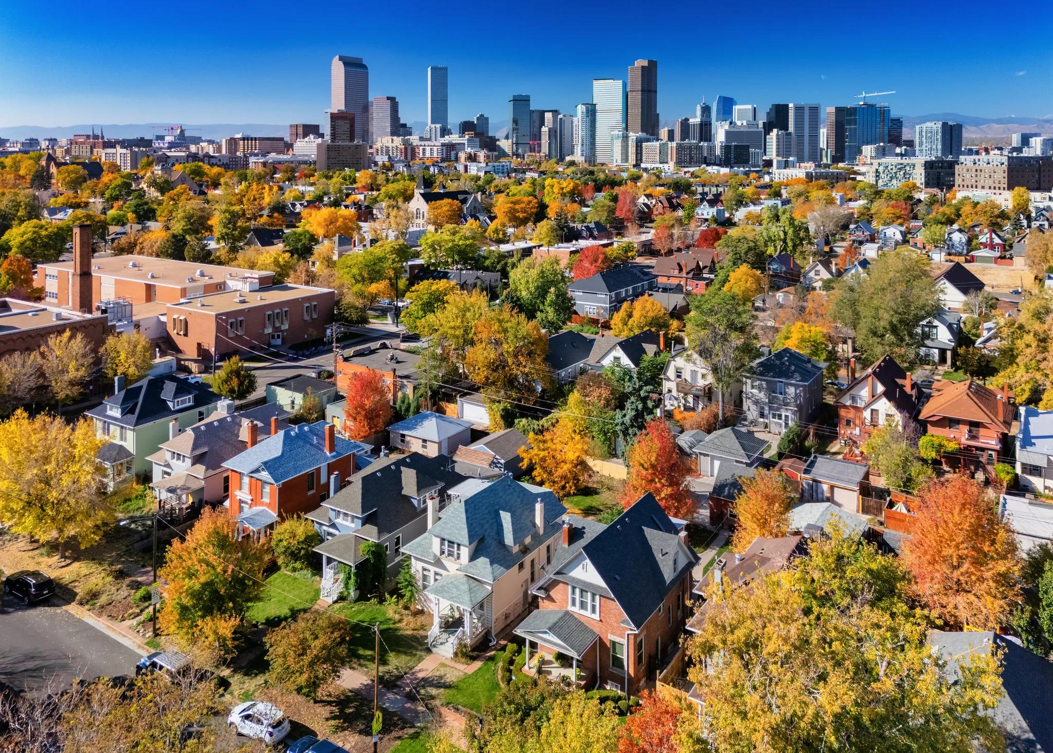 Aerial view of a residential neighborhood with fall foliage in the foreground and a downtown city skyline in the background.