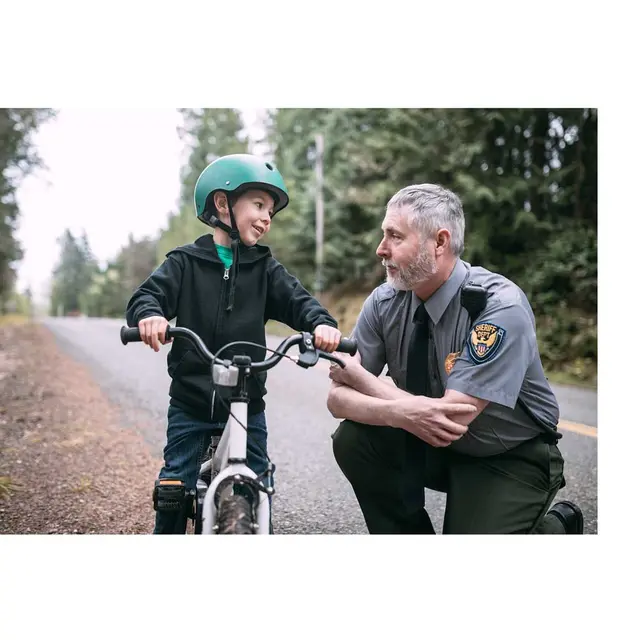 Public safety law enforcement officer kneeling low to speak with a child on a bike.