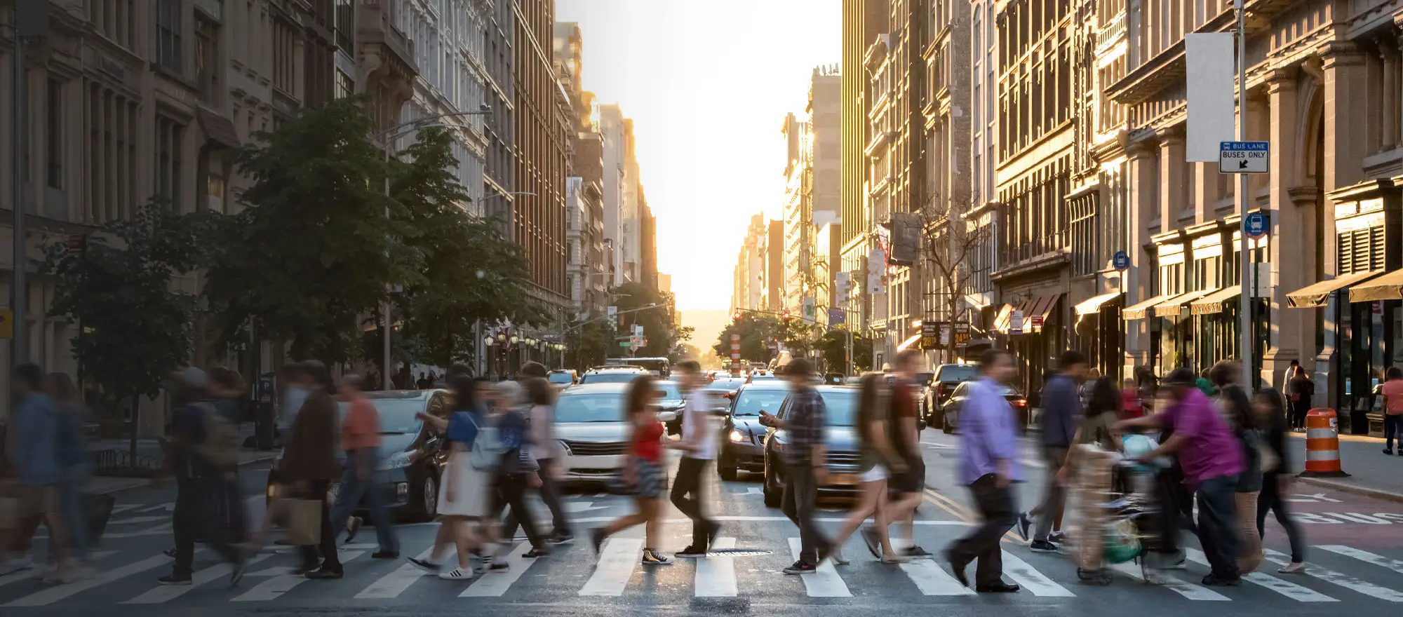 An image showing a vibrant community. A bright sunset in the background and people crossing a busy street at a crosswalk in the foreground.