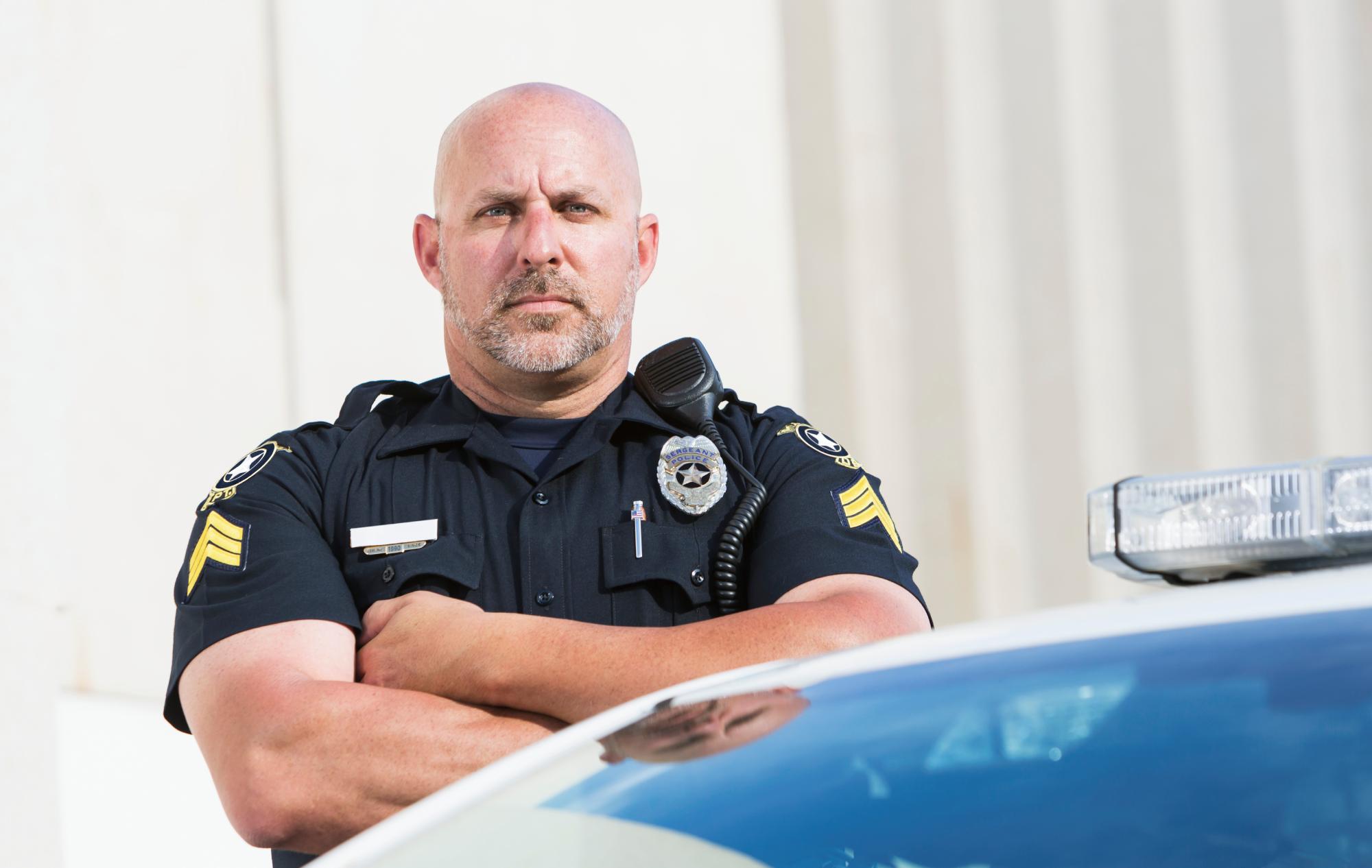 A trusted police officer standing, arms folded, in front of his police cruiser.